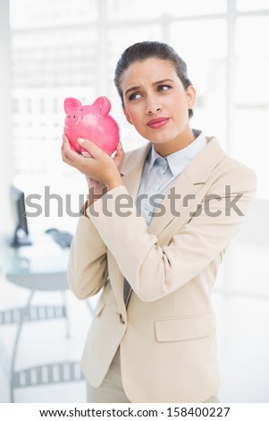 Frowning smart brown haired businesswoman shaking a piggy bank in bright office