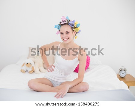 Cheerful natural brown haired woman in hair curlers sitting on her bed in bright bedroom