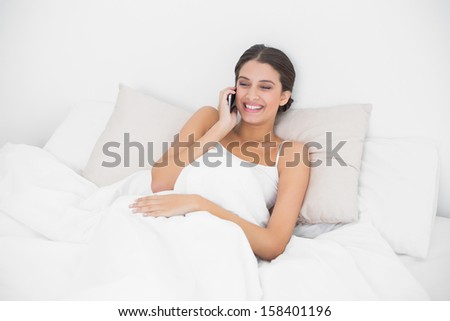 Laughing young brown haired model in white pajamas calling with her mobile phone in bright bedroom