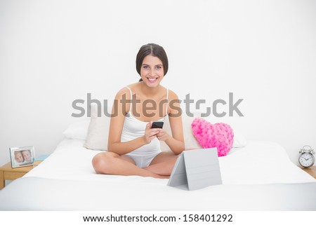 Content young brown haired model in white pajamas using a mobile phone in bright bedroom