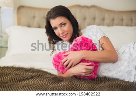 Peaceful pretty brown haired woman hugging a heart shaped pillow in a chic bedroom