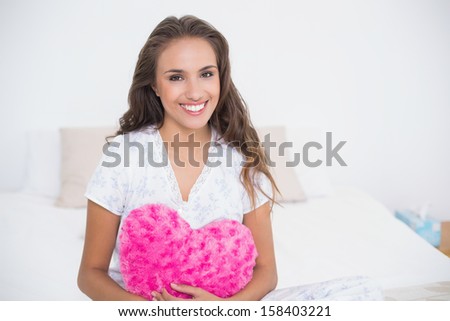 Smiling attractive woman holding heart pillow in bright bedroom