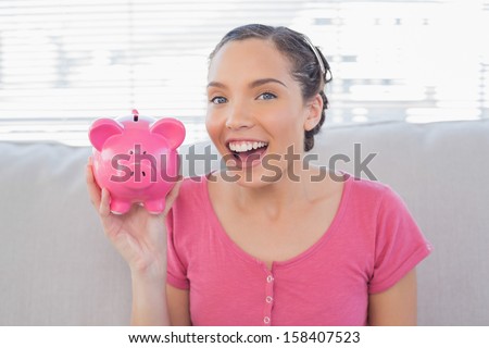 Happy woman showing piggy bank while sitting on sofa and looking at camera