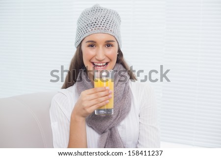 Pretty brunette with winter hat on holding glass of orange juice sitting on cosy sofa