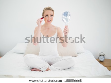 Natural happy blonde holding mirror and applying mascara in bright bedroom