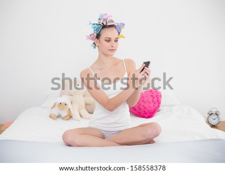Stern natural brown haired woman in hair curlers using her mobile phone in bright bedroom