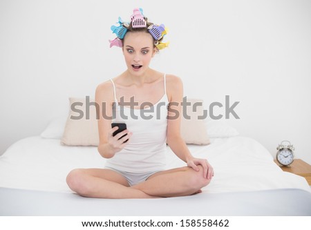 Astonished natural brown haired woman in hair curlers looking at her mobile phone in bright bedroom