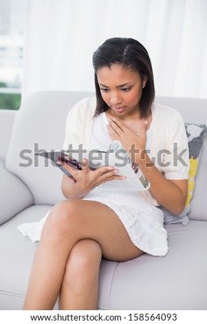 Surprised young dark haired woman in white clothes reading a magazine in a living room