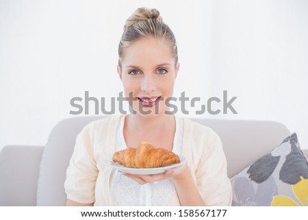 Happy fresh model holding croissant sitting on sofa in bright living room