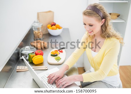 Smiling cute blonde typing on her laptop in bright kitchen