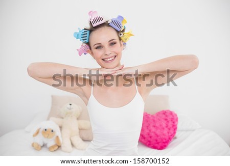 Joyful natural brown haired woman in hair curlers posing looking at camera in bright bedroom