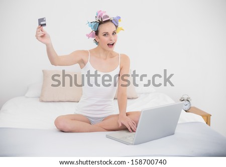 Excited natural brown haired woman in hair curlers shopping online with her laptop in bright bedroom