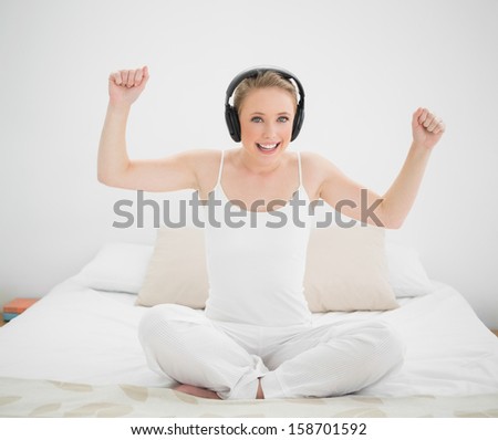 Natural smiling blonde listening to music and arms in the air in bright bedroom