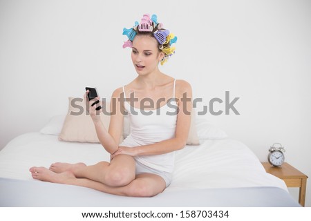 Amazed natural brown haired woman in hair curlers looking at her mobile phone in bright bedroom