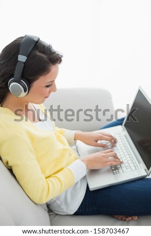 Calm casual brunette in yellow cardigan listening to music while using a laptop sitting on a couch in bright living room