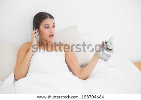 Surprised young brown haired model in white pajamas on the phone looking at her alarm clock in bright bedroom