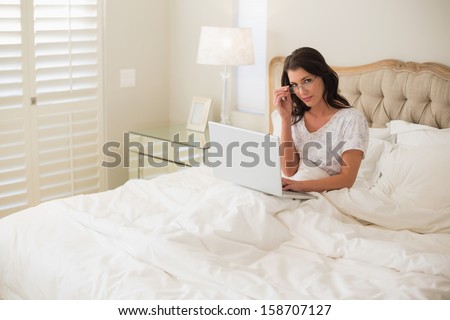 Stern pretty brown haired woman using a laptop in a bright bedroom