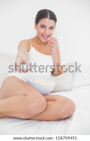 Concentrated young brown haired model in white pajamas eating popcorn while watching tv in bright bedroom