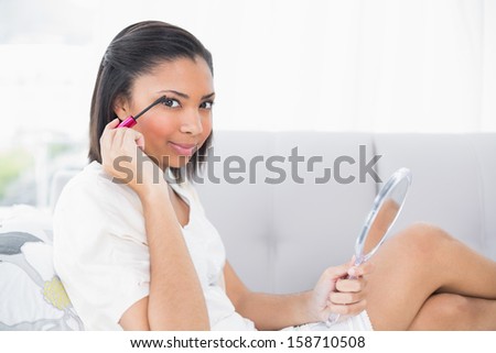 Playful young dark haired woman in white clothes applying mascara in a living room