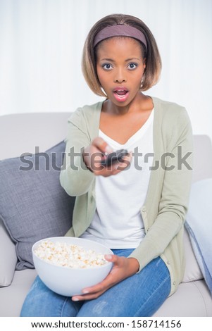 Shocked woman sitting on sofa changing tv channel while holding popcorn in living room