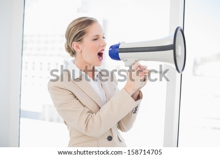 Angry blonde businesswoman in bright office shouting in megaphone
