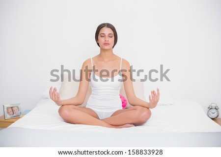 Serene young brown haired model in white pajamas practicing yoga in bright bedroom