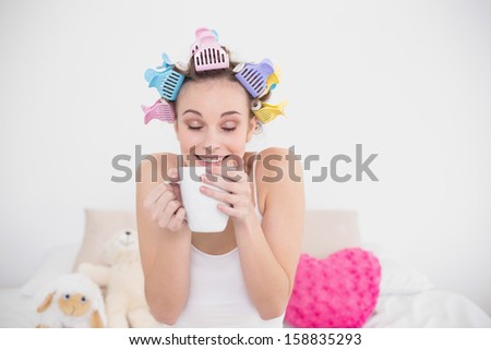 Funny natural brown haired woman in hair curlers enjoying coffee smell in bright bedroom