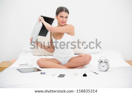Furious young brown haired model in white pajamas throwing her laptop in bright bedroom