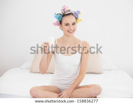 Pretty natural brown haired woman in hair curlers holding a glass of milk in bright bedroom