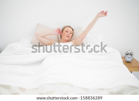 Natural yawning blonde lying in bed with closed eyes and stretched arms in bright bedroom