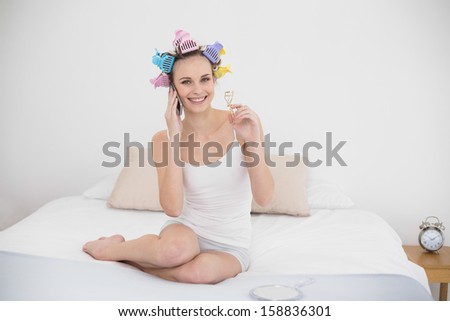 Content natural brown haired woman in hair curlers making a phone call and holding an eyelash curler in bright bedroom