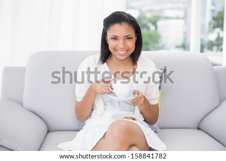 Pleased young dark haired woman in white clothes enjoying coffee and cookies in a living room