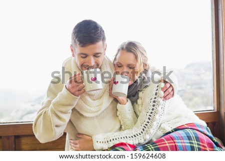 Loving young couple in winter clothing drinking coffee against cabin window