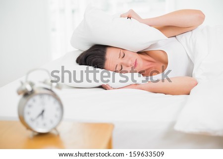 Young woman covering ears with pillow in bed and alarm clock on side table