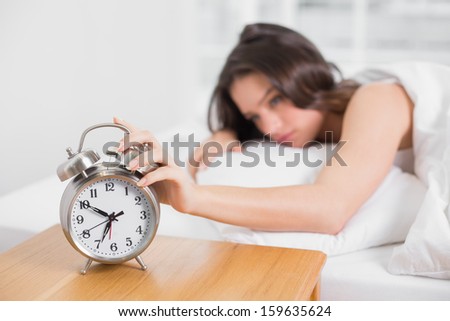 Sleepy young woman in bed extending hand to alarm clock at home