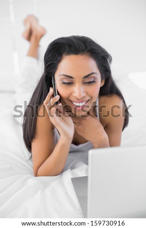 Happy natural woman phoning while using her notebook and lying on her bed