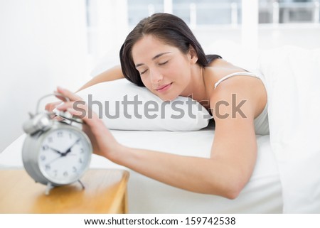 Young woman in bed extending hand to alarm clock at home