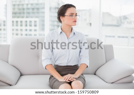 Serious well dressed young woman sitting on sofa and looking away at bright home