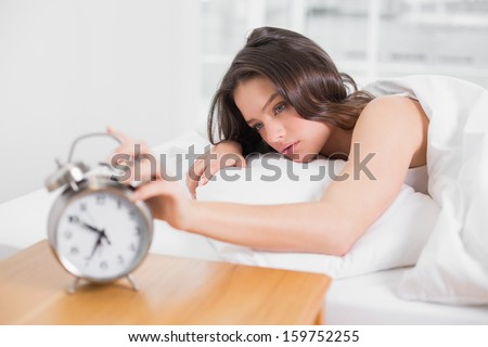 Sleepy young woman in bed extending hand to alarm clock at home