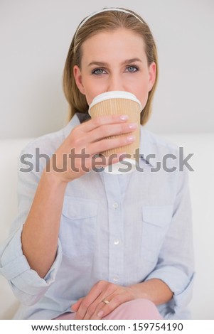 Calm casual blonde sitting on couch drinking from disposable cup in bright living room