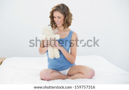 Pretty sweet woman holding her white teddy bear looking at it sitting on her bed in the bedroom
