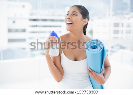 Laughing toned brunette holding sports bottle and exercise mat in bright room