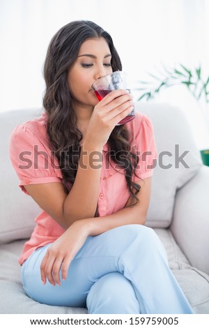 Calm cute brunette sitting on couch drinking glass of juice in bright living room