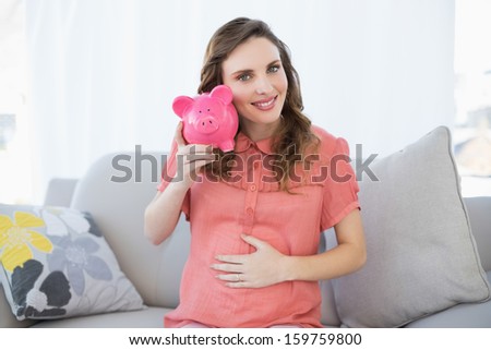Content pregnant woman shaking pink piggy bank sitting on couch in living room