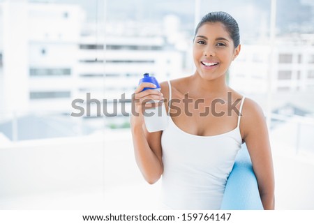 Happy toned brunette holding sports bottle and exercise mat in bright room