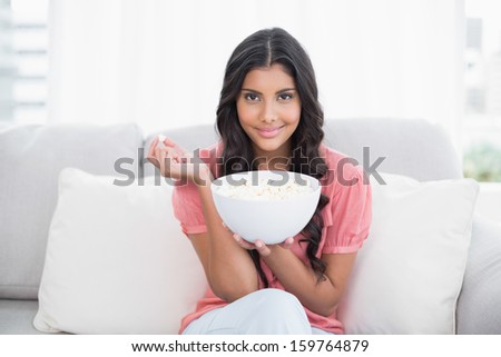 Content cute brunette sitting on couch holding popcorn bowl in bright living room