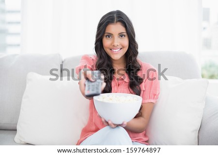 Smiling cute brunette sitting on couch holding popcorn bowl in bright living room