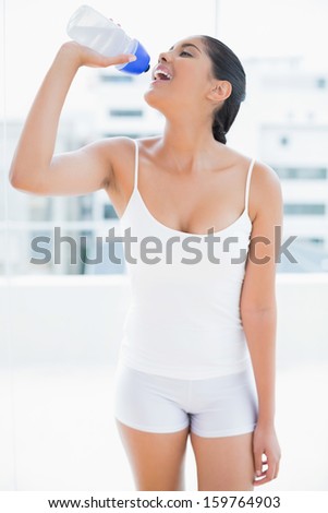Happy toned brunette drinking from sports bottle in bright room