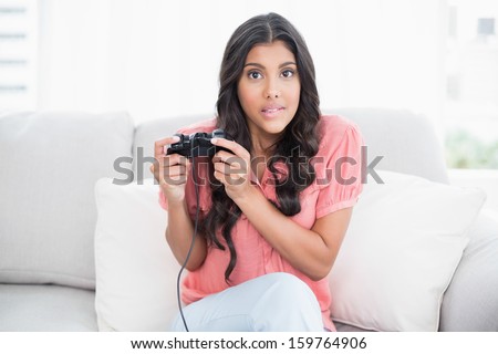Shocked cute brunette sitting on couch holding controller in bright living room