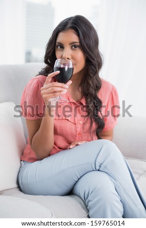 Happy cute brunette sitting on couch drinking red wine in bright living room